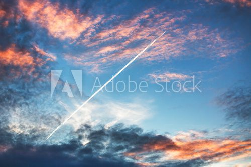 Fototapeta Airplane flies in sunset dramatic clouds and leaving trail