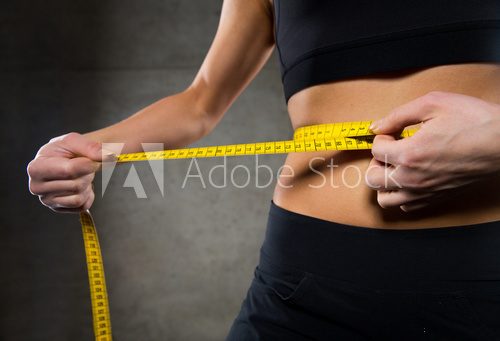 Fototapeta close up of woman measuring waist by tape in gym