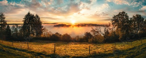 Fototapeta Magnificent sunrise over rural idyll. A colorful panorama with romantic, dramatic sky, sunlit fog in the valley and tree silhouettes