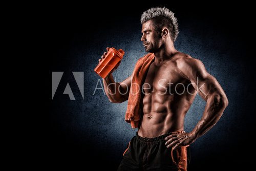 Fototapeta Young adult man drinking protein shake in gym. Black background.